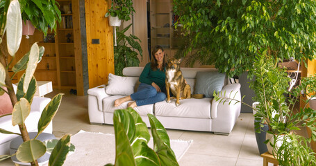 PORTRAIT: Young woman and her brown shepherd dog relax together on a white couch in a modern living room surrounded by thriving houseplants. They share a peaceful, joyful moment on a quiet day at home