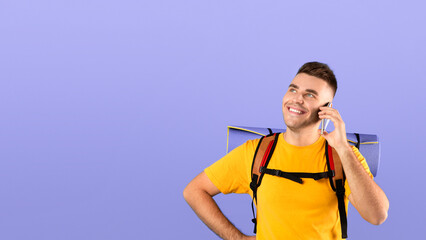 Naklejka premium Attractive millennial guy with tourist gears talking on smartphone over blue studio background. Happy young man having friendly conversation on mobile phone during his travels