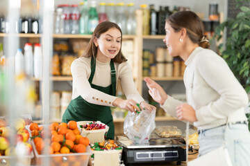 Grocery store selling area. Female employee sells candies to woman, puts them in bag, puts them on scale and weighs sweets. Brunette European girl buys candies by weight.
