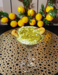 Pistachio dessert in martini glass with ladyfingers. Elegant pistachio mousse served in a martini glass, topped with crushed nuts and ladyfingers, set against lemon-themed decor.