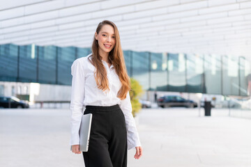 Fototapeta premium Smiling businesswoman with long hair standing with closed laptop. Young woman wears white shirt and dark pants. Modern office building is in background