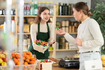 Female seller in green apron holds out basket with hanging sweets and invites girl buyer to familiarize with product. Customer of store takes candy in bright package and examines sweetness.