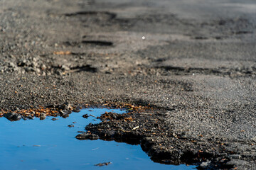 Standing water puddle on worn asphalt surface, rocks and debris visible along water's edge, close up