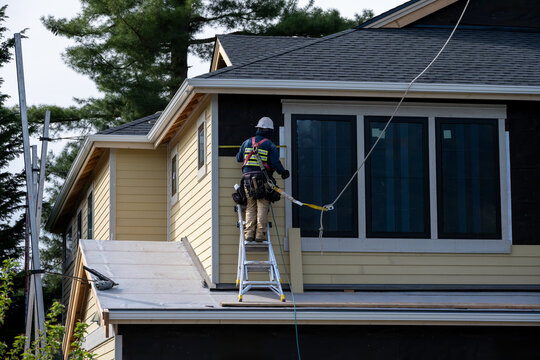 Carpenter on ladder installing wood siding 2nd floor exterior wall on new home construction, safety harness, hard hat, and toolbelt
