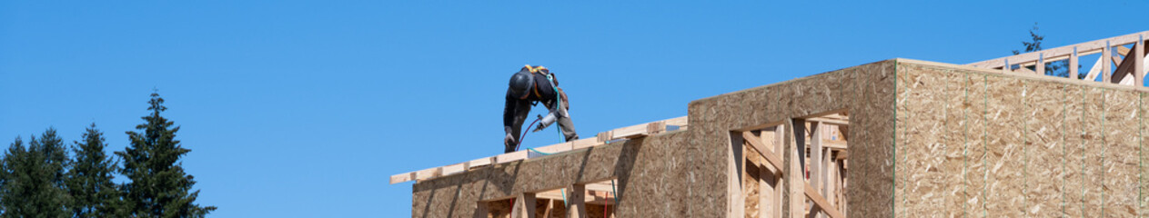 Framing contractor on roof of new home construction installing roof trusses with nail gun, sunny...