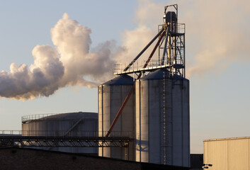 Industrial Plant with Grain Silos and Emission Against Sky at Dusk Producing Vapor Plumes in Evening Light