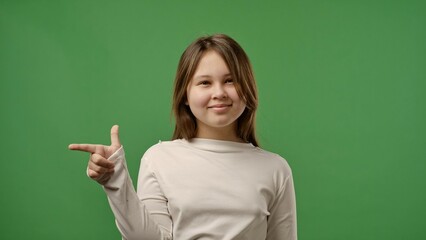 Teenage girl posing at camera pointing finger at side, positive expression, isolated on chroma key green screen studio background mockup.