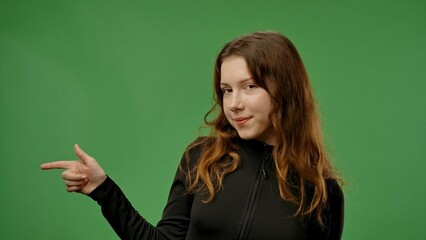 Teenage girl posing at camera pointing finger at side, positive expression, isolated on chroma key green screen studio background mockup.