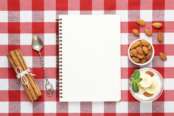 Blank recipe book, banana dessert, spoon and ingredients on checkered tablecloth, top view. Mockup...