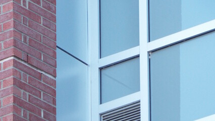 A close-up shows a building's exterior where a traditional red brick wall meets a modern glass facade with white frames reflecting a clear, light blue sky. 