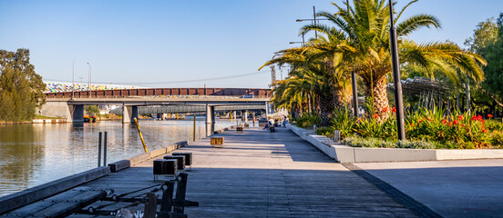 Footscray Wharf in Melbourne, Australia, with palm trees, landscaped gardens, and a pedestrian boardwalk beside the Maribyrnong River—riverside regeneration, public recreation, and modern urban design