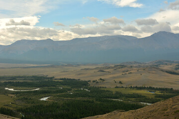 Panoramic view of a wide, endless steppe with a meandering river at the foot of snow-capped mountain peaks on a cloudy summer morning.