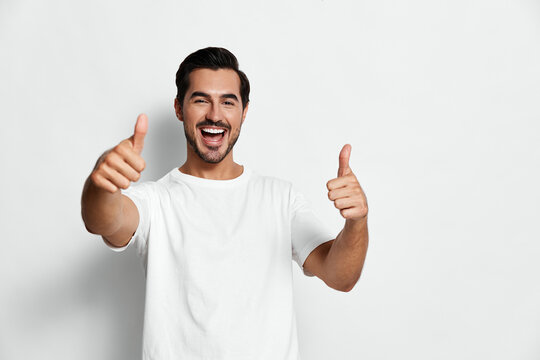 Full body smiling joyful young man gives thumbs up, wear casual white t-shirt, look at camera, isolated on clean solid light background studio portrait. People happiness concept