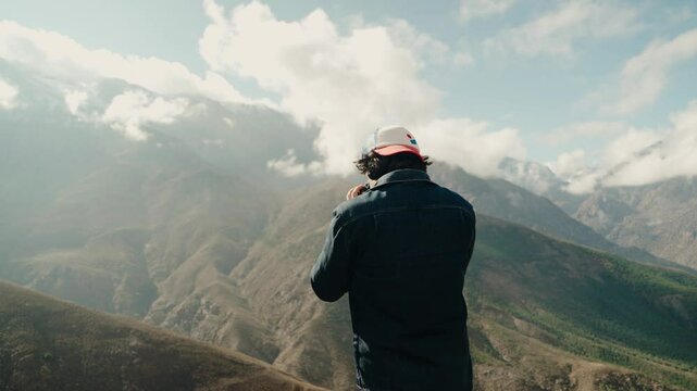 A man wearing a baseball cap and denim jacket takes in the majestic mountain view in Morocco. The rugged landscape stretches out before him, with clouds drifting over the peaks.