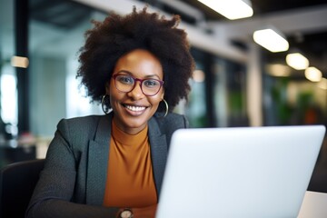 A smiling businesswoman with an afro hairstyle, wearing glasses, sitting at a desk with a laptop in an office setting.