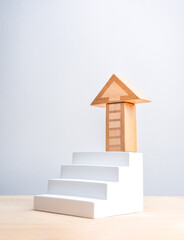 Wood block arranged as arrow on top of white geometric stair on wood table and vertical grey background. Business market growth, mission, goal and success process and economic improvement concepts.