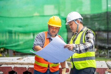 Two Construction Workers Discussing Blueprints on a Construction Site with Green Safety Nets and Hard Hats in Bright Daylight