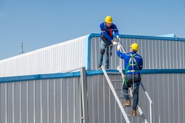 Two construction workers wearing safety gear climb up a ladder to the roof of a building