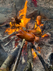 Sausages and bacon roasting on wooden sticks over an open campfire in a forest, with visible flames, rocks, and burning wood.