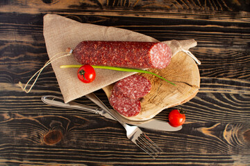 A piece of smoked sausage, cutlery and cherry tomatoes in close-up on a dark wooden background