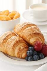 Two flaky croissants on a white plate with fresh raspberries and blueberries. A bowl of orange slices and a cup of coffee are in the background