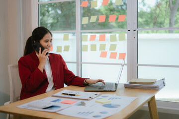 Asian businesswoman using smartphone and working on laptop while sitting at desk in home office.