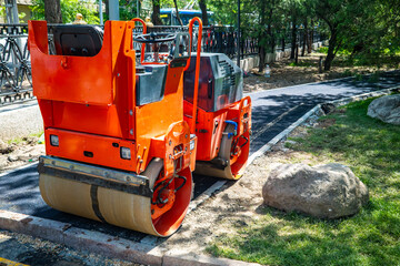 orange asphalt roller parked at roadside during construction, compact road compaction machine with dual steel drums on freshly paved street near park fence