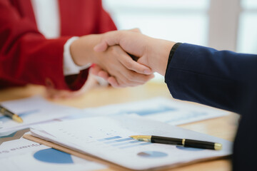 Close-up businessman handshake over documents showing accounting and financial graphs and charts on desk in office space.