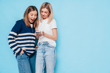 Two young beautiful smiling brunette hipster female in trendy summer clothes. Carefree women posing near blue wall. Positive models having fun. Cheerful and happy. Holding smartphone. Using apps
