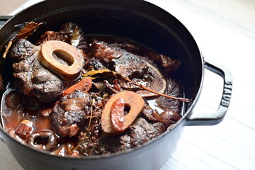 Close-up of a rustic gourmet dish featuring slow-braised beef shank with bone (osso buco style), served on black pot ,white background