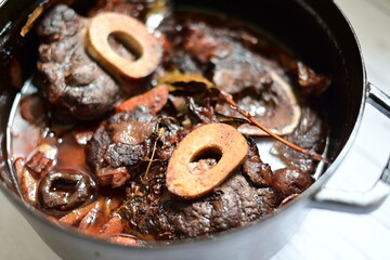 Close-up of a rustic gourmet dish featuring slow-braised beef shank with bone (osso buco style), served on black pot ,white background
