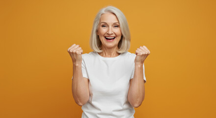 Senior woman in white t-shirt expressing excitement with raised fists against a mustard yellow backdrop. Celebration pose of a mature female professional in a studio setting.