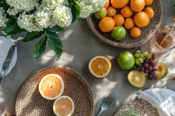 Summer Fruit Feast on Woven Table. A summer table setting featuring fresh fruits, flowers, and woven placemats, perfect for a sunny day. Focus is on colorful arrangement of oranges, limes, and grapes