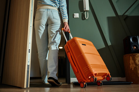 Woman exits from apartment with orange suitcase. Person leaving home with suitcase. Traveler existing door with luggage