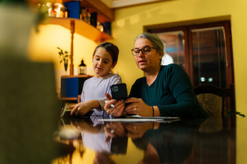 Fototapeta premium Grandmother and granddaughter sitting together at the dining table, engaging with a smartphone and textbook for homework, enjoying a moment of learning and connection on the polished surface