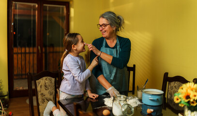 Grandmother wearing apron putting flour on granddaughter's nose while kneading dough, making a...
