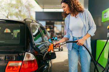 Young woman refueling her car at a gas station on a sunny day, holding the fuel pump and filling...