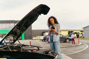 Young woman is standing next to her broken car with open hood in parking lot, carefully reading...
