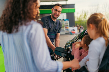 Fototapeta premium Friendly gas station attendant refueling a family car while chatting with the mother and her two daughters sitting comfortably inside the vehicle during their summer road trip
