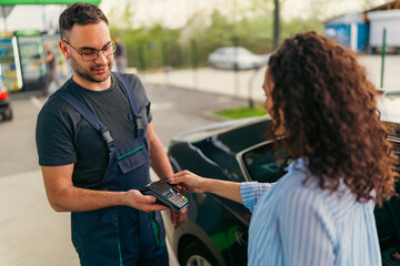 Fototapeta premium Customer making a contactless payment using her smartphone at a gas station, while a mechanic holds the pos terminal, providing convenient and modern transaction
