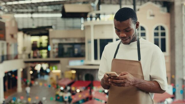 Portrait of African American man with smile standing with smartphone near restaurant. Cheerful male waiter checks social media during break. Guy service worker in trade mall cafe