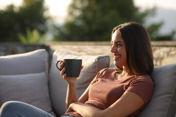 Happy woman drinking coffee sitting on a sofa in a terrace