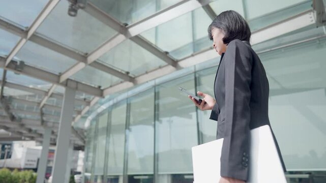 A woman is walking with a white folder and a cell phone. She is looking at her phone