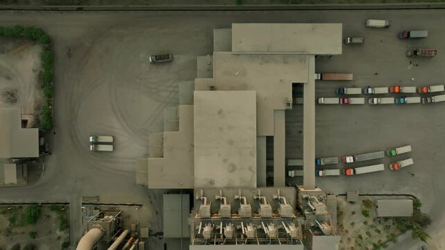 Top-down real time view of a cement factory with parallel truck lanes and industrial structures framed by paved lots and dust trails, aerial view, drone shot