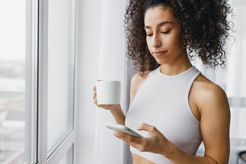 Side view cropped image of good-looking black female in tank-top standing next to window with cup...