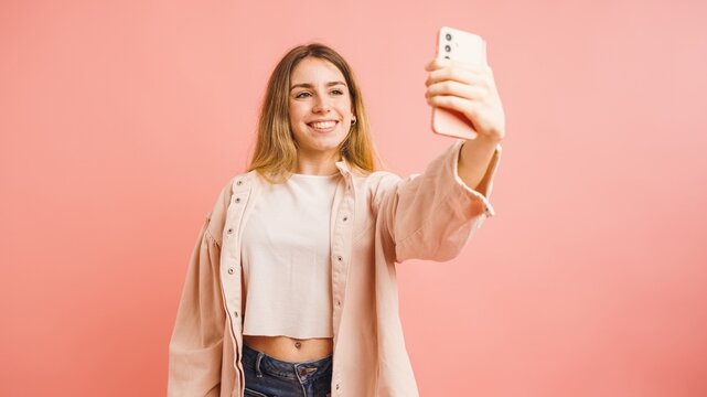 Young woman taking selfie with smartphone on pink background