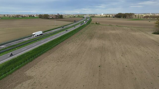 Vehicles including a cargo truck travel along the A1 motorway across the flat agricultural land near Piacenza, with scattered rural buildings and tilled fields extending into the horizon, slow motion