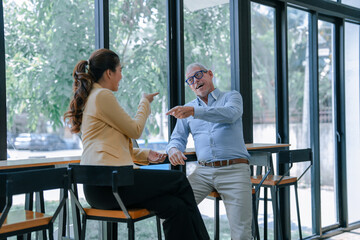Two colleagues are sitting at a table in the office cafeteria, pointing fingers at each other and...