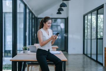 Young asian businesswoman sitting on desk and using smartphone in modern office, she's smiling and...