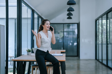 Fototapeta premium Asian businesswoman sitting at a modern desk in a bright office, talking on her phone while gesturing with her hand, smiling confidently and looking away, exuding professionalism and success